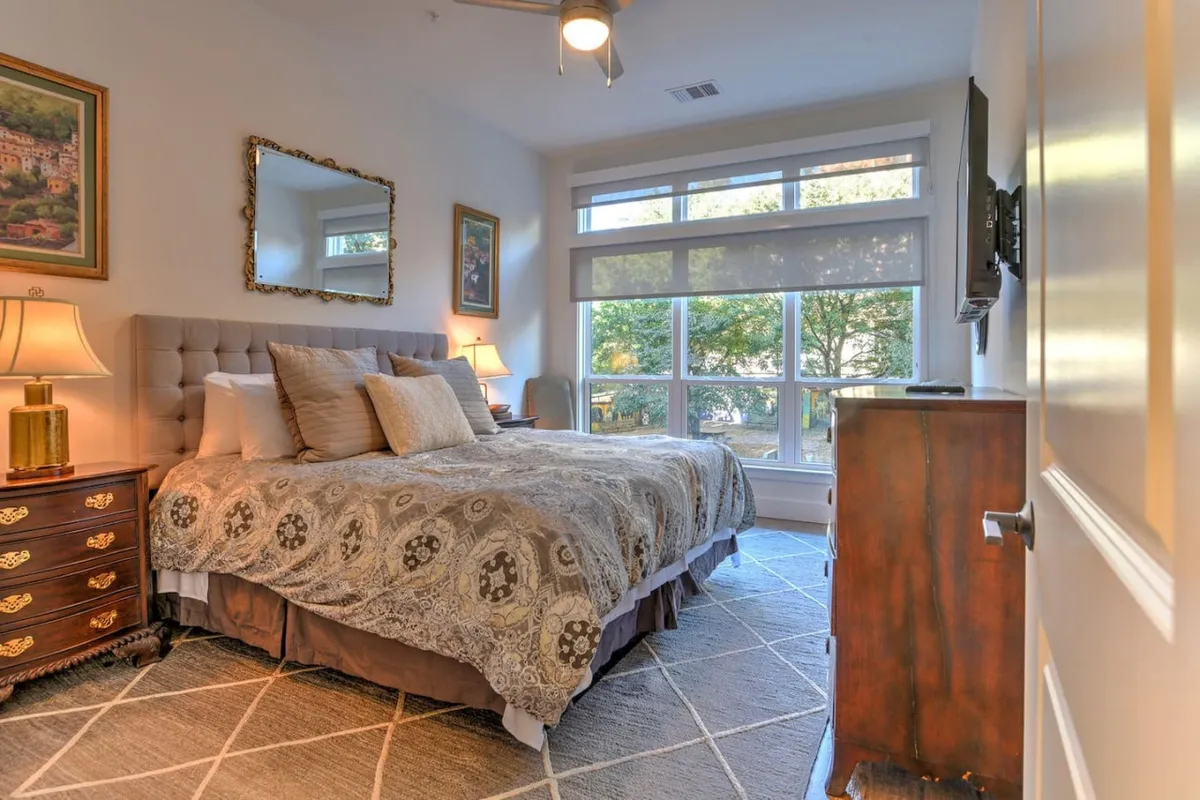 A calm bedroom scene with a plush bed, windows that allow plenty of natural light, and a nightstand. The windows provide a glimpse of the outside world and suggest a restful, hotel-style atmosphere for unwinding after a day out.