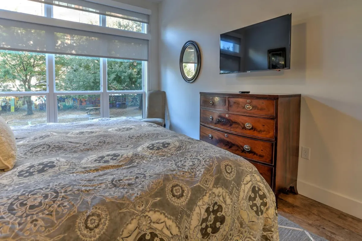 A spacious and serene bedroom featuring a king-sized bed adorned with patterned luxury bedding and plush pillows. The room is accented by a wooden dresser, a wall-mounted TV, a round mirror, and large windows offering ample natural light and an inviting view outside. The overall atmosphere is cozy and restorative, perfect for relaxation and unwinding.