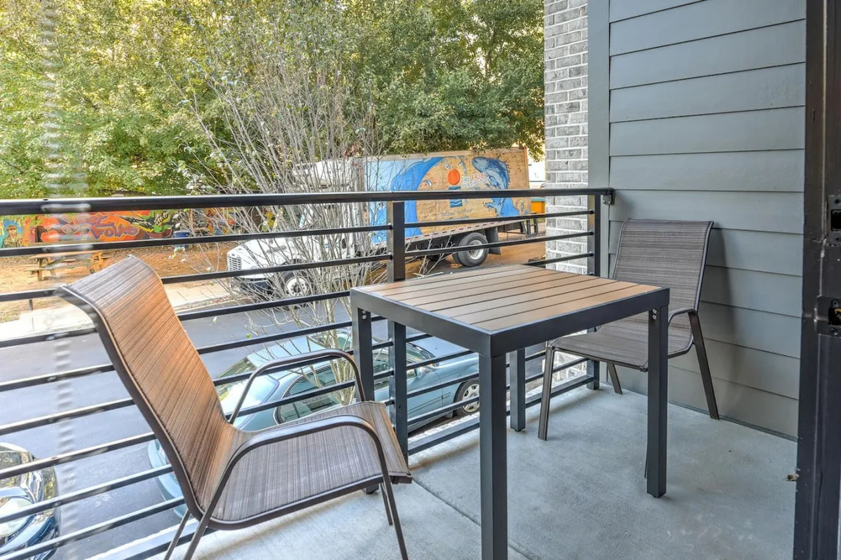A charming balcony setup with two chairs and a small table, overlooking a street lined with trees. The mood is laid-back and perfect for sipping a morning coffee, watching the world go by, or sharing a sunset toast, as described in the narrative.