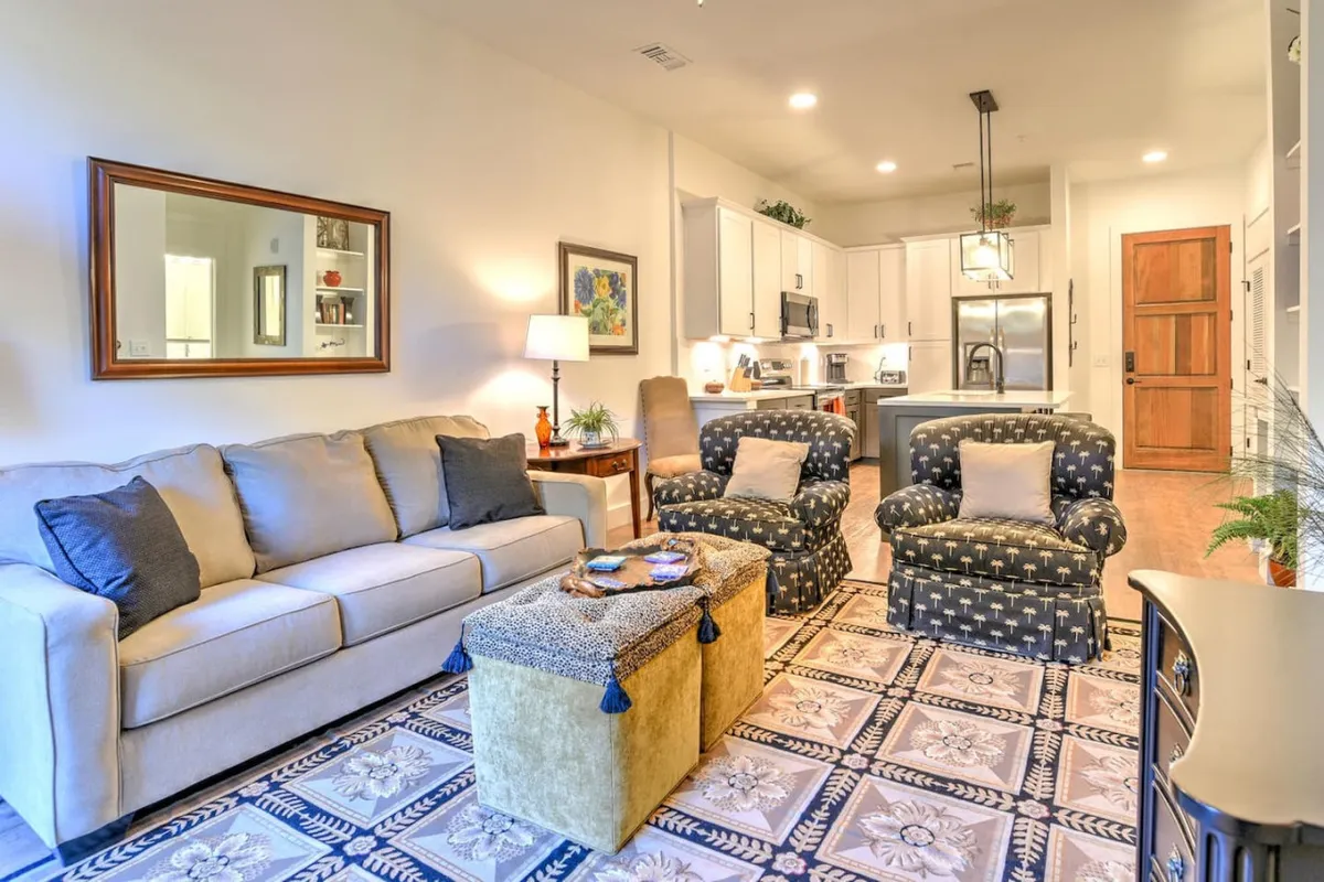 A bright, open-concept living space featuring a comfortable sofa, accent chairs, a central coffee table, a stylish rug, and a view toward a contemporary kitchen area. The layout encourages relaxation, conversation, or smooth transitions from one activity to another.