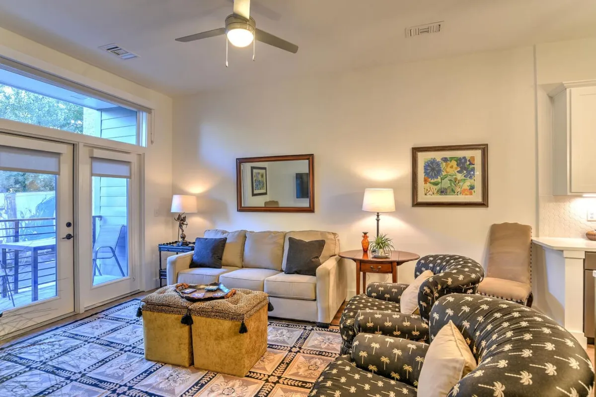 The image showcases a stylish, light-filled living area with a plush sofa, inviting armchair, and a modern coffee table, thoughtfully accented by decorative touches. Floor-to-ceiling windows bathe the room in warm natural light, hinting at a beautiful sunrise and views of vibrant downtown Asheville. The cozy ambiance is enhanced by soft lighting and comfortable seating, creating the perfect spot for relaxing or socializing.