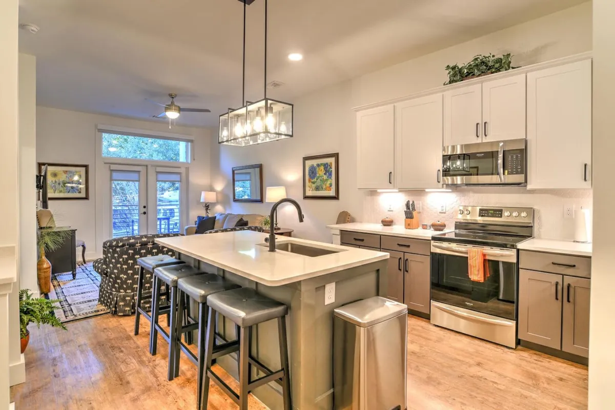 A spacious, welcoming kitchen area featuring a large kitchen island with modern seating, gleaming stainless steel appliances, and a comfortable seating area visible in the background. The setting exudes an upscale, hotel-like ambiance while remaining inviting and functional for cooking or entertaining.