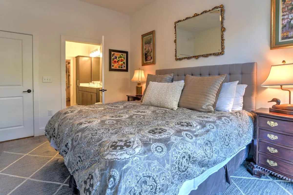 A cozy bedroom scene with a plush bed dressed in luxury bedding, a decorative mirror, a nightstand with a softly glowing lamp, and tasteful wall art that elevates the room's ambiance. Soft lighting and thoughtful decor add warmth and elegance.