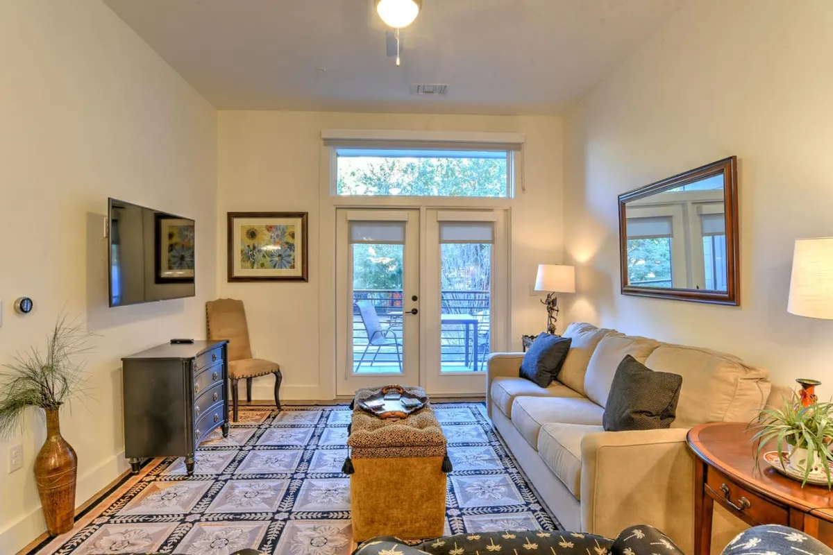 This image features a comfortable living room with a sofa, coffee table, TV, and double doors leading outside. Sunlight pours into the space, and the view through the double doors hints at a private outdoor area or balcony, connecting the interior with the outdoors.